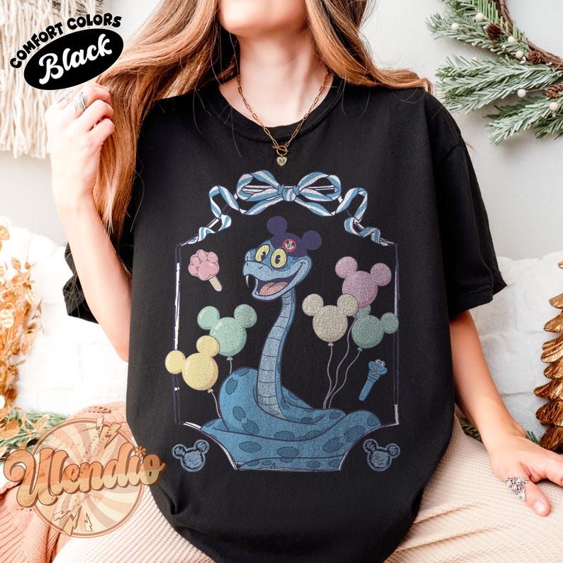 Puede incluir: Camiseta negra con una ilustraci&oacute;n de dibujos animados de una serpiente azul con un sombrero tem&aacute;tico de Mickey Mouse y globos. La serpiente sonr&iacute;e y est&aacute; rodeada de globos de colores. El texto "Comfort Colors Black" est&aacute; en un &oacute;valo negro.
