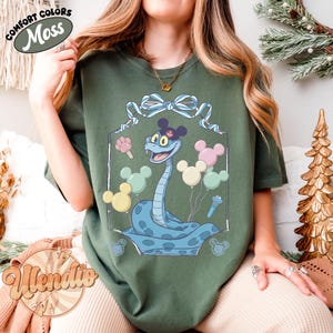 Puede incluir: Camiseta verde musgo con una serpiente de dibujos animados con un sombrero de Mickey Mouse y globos. La serpiente es azul claro con un patr&oacute;n azul oscuro. La camiseta tiene las palabras "Comfort Colors" y "Moss".