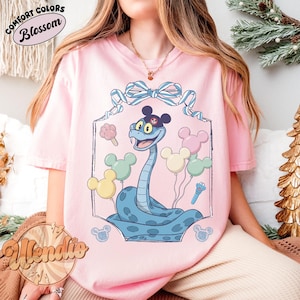 Puede incluir: Camiseta rosa claro con una ilustraci&oacute;n de una serpiente de dibujos animados. La serpiente azul lleva un sombrero negro y est&aacute; rodeada de globos de Mickey Mouse en colores pastel. La camiseta tiene la inscripci&oacute;n "Comfort Colors Blossom".