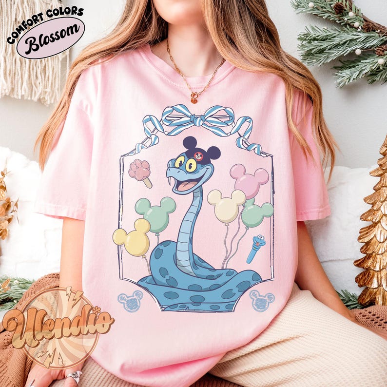 Puede incluir: Camiseta rosa claro con una ilustraci&oacute;n de una serpiente de dibujos animados. La serpiente azul lleva un sombrero negro y est&aacute; rodeada de globos de Mickey Mouse en colores pastel. La camiseta tiene la inscripci&oacute;n "Comfort Colors Blossom".