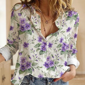 May include: A white button-down shirt with a vibrant floral pattern of purple violets and green leaves. The long-sleeved shirt has a relaxed fit and is made of a lightweight fabric. The shirt is unbuttoned at the top, revealing a glimpse of the wearer's neckline.
