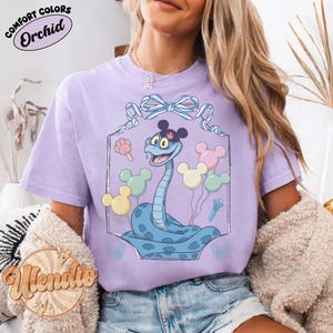 Puede incluir: Camiseta lila con un dise&ntilde;o de serpiente de dibujos animados. La serpiente azul lleva un sombrero negro y est&aacute; rodeada de globos y helados. La camiseta tiene la inscripci&oacute;n "Comfort Colors Orchid".