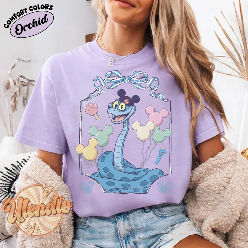Puede incluir: Camiseta lila con un dise&ntilde;o de serpiente de dibujos animados. La serpiente azul lleva un sombrero negro y est&aacute; rodeada de globos y helados. La camiseta tiene la inscripci&oacute;n "Comfort Colors Orchid".