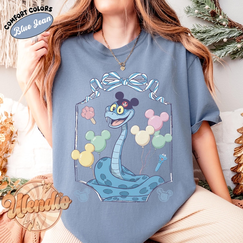 Puede incluir: Camiseta azul vaquero con una serpiente de dibujos animados con un sombrero de Mickey Mouse y globos. La serpiente est&aacute; rodeada por un marco decorativo con un lazo en la parte superior. La camiseta tiene el texto "Comfort Colors" y "Blue Jean".