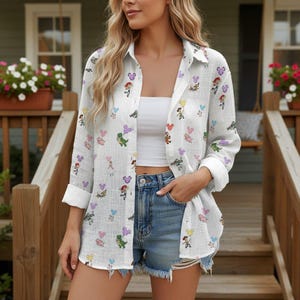 May include: White button-down shirt with a colorful cartoon character pattern. The shirt has long sleeves, rolled up at the elbows, and is paired with denim shorts. The shirt features a classic collar and a relaxed fit.