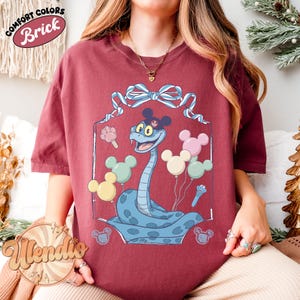 Puede incluir: Camiseta color ladrillo con una serpiente de dibujos animados con orejas y globos de Mickey Mouse. La serpiente es azul claro con un contorno azul oscuro. La camiseta tiene las palabras "Comfort Colors" y "Brick" en un dise&ntilde;o circular.
