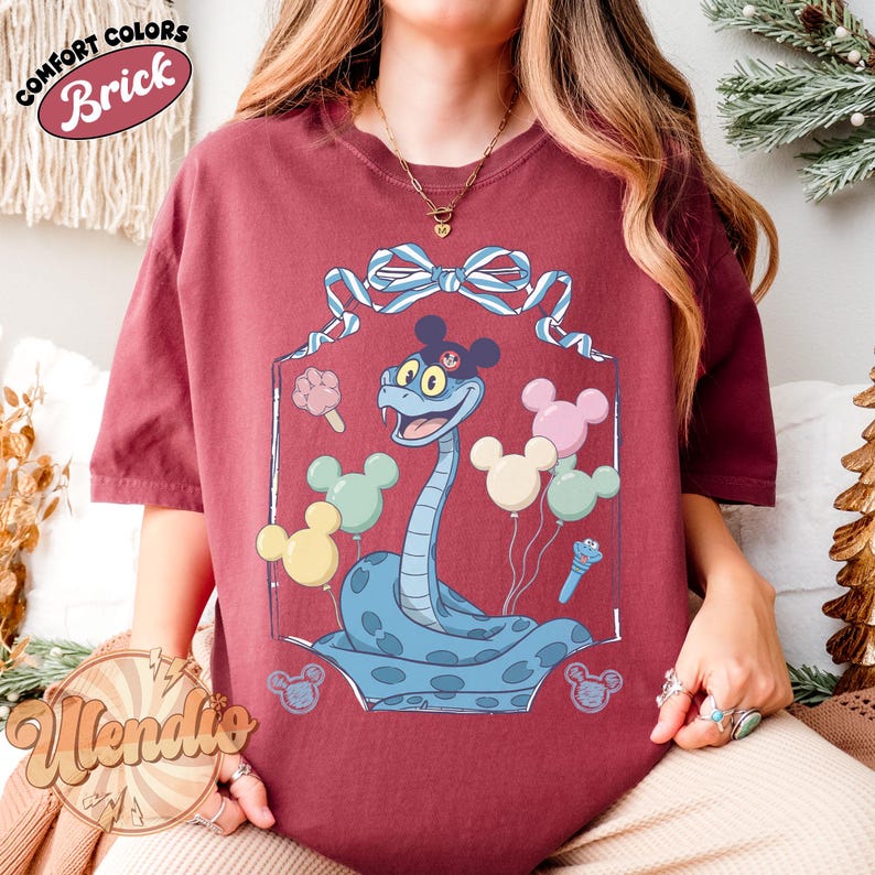 Puede incluir: Camiseta color ladrillo con una serpiente de dibujos animados con orejas y globos de Mickey Mouse. La serpiente es azul claro con un contorno azul oscuro. La camiseta tiene las palabras "Comfort Colors" y "Brick" en un dise&ntilde;o circular.