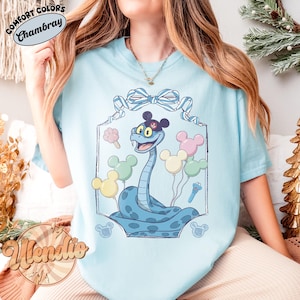 Puede incluir: Camiseta azul claro con una ilustraci&oacute;n de dibujos animados de una serpiente azul con sombrero y globos de Mickey Mouse. La camiseta tiene el texto "Comfort Colors Chambray" en la parte superior y "Wendie" en la inferior. El dise&ntilde;o est&aacute; enmarcado.