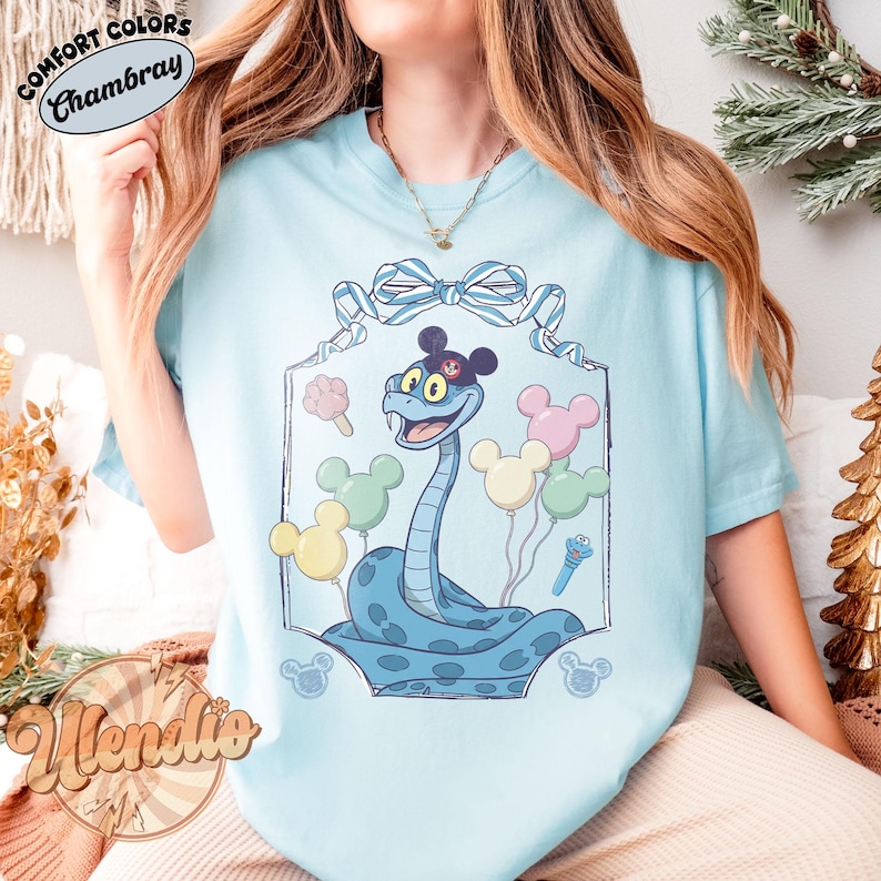 Puede incluir: Camiseta azul claro con una ilustraci&oacute;n de dibujos animados de una serpiente azul con sombrero y globos de Mickey Mouse. La camiseta tiene el texto "Comfort Colors Chambray" en la parte superior y "Wendie" en la inferior. El dise&ntilde;o est&aacute; enmarcado.