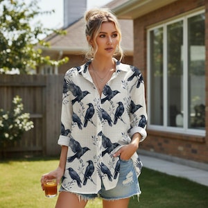 May include: A long-sleeved button-down shirt in an off-white color featuring a repeating pattern of black crows perched on thin branches. The shirt has a classic collar, a front button placket, and cuffs that are rolled up to the mid-forearm.