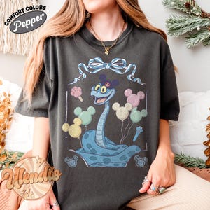 Puede incluir: Camiseta gris oscuro con un dise&ntilde;o de serpiente azul de dibujos animados, con orejas de Mickey Mouse y globos y helados. La camiseta tiene el texto "Comfort Colors Pepper" y "Wendie" en un logotipo circular.
