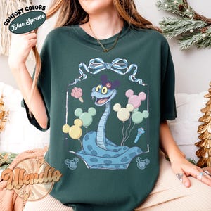 Puede incluir: Camiseta verde oscuro con una serpiente de dibujos animados con orejas de Mickey Mouse, rodeada de globos y helados con forma de Mickey Mouse. La camiseta tiene el texto "Comfort Colors Blue Spruce" y "Allendio".