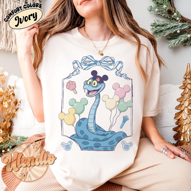 Puede incluir: Camiseta de color marfil con un dise&ntilde;o de serpiente de dibujos animados. La serpiente es de color azul claro con manchas azul oscuro y orejas de Mickey Mouse. La camiseta tiene las palabras "Comfort Colors Ivory" en la parte superior y "Wendie's" en la parte inferior.