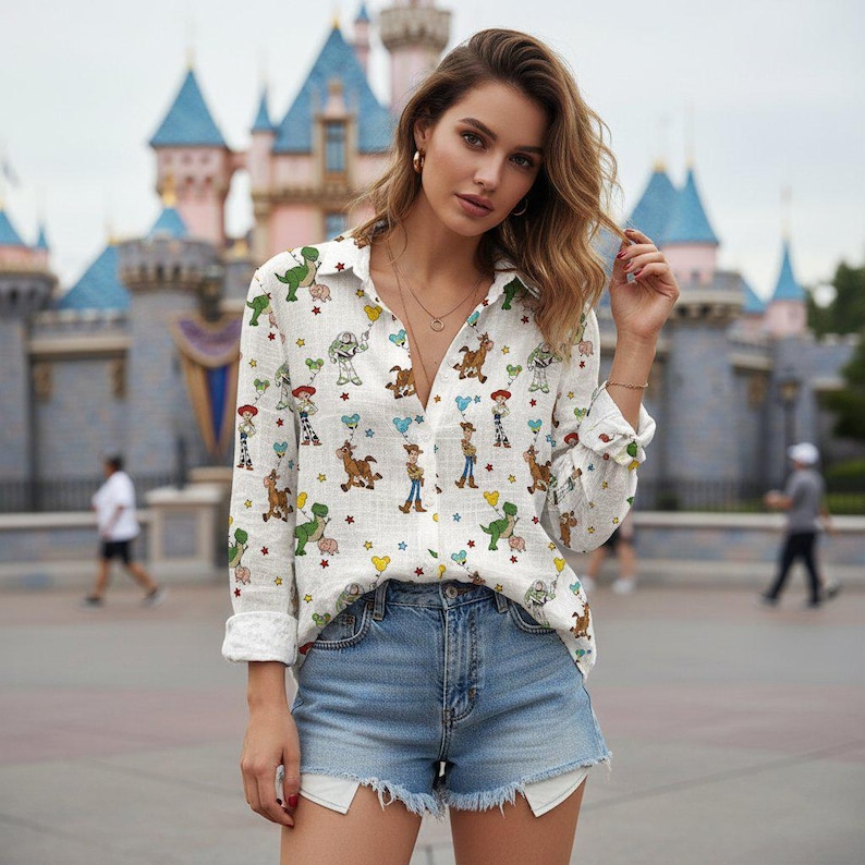 May include: White button-down shirt with a colorful pattern of cartoon characters, paired with denim shorts. The shirt features long sleeves and a classic collar. The background includes a castle and people.