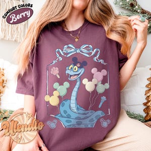 Puede incluir: Camiseta color bayas con una serpiente de dibujos animados con orejas de Mickey Mouse y globos. La serpiente es azul claro con manchas azul oscuro. La camiseta tiene las palabras "Comfort Colors" y "Berry" en un dise&ntilde;o circular.