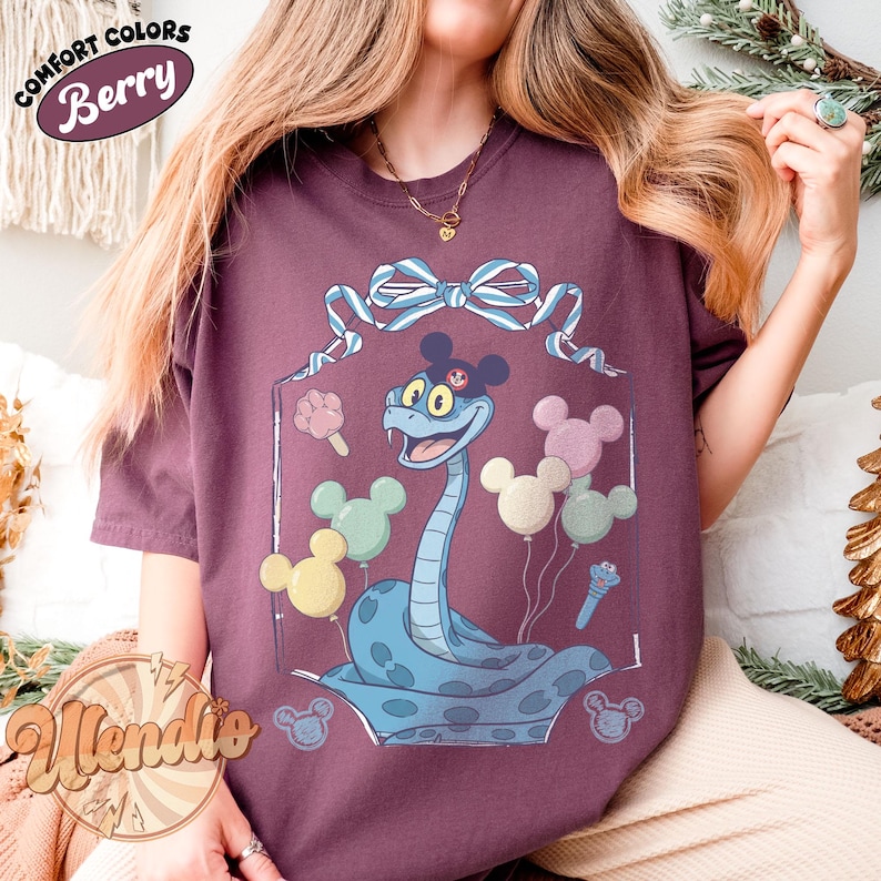 Puede incluir: Camiseta color bayas con una serpiente de dibujos animados con orejas de Mickey Mouse y globos. La serpiente es azul claro con manchas azul oscuro. La camiseta tiene las palabras "Comfort Colors" y "Berry" en un dise&ntilde;o circular.