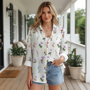 May include: A white button-down shirt with a colorful cartoon character pattern. The shirt features rolled-up sleeves and is paired with denim shorts. The pattern includes cartoon characters, balloons, and other playful elements.