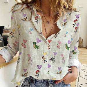 May include: A light green button-down shirt with a repeating pattern of cartoon characters and Mickey Mouse-shaped balloons. The long-sleeved shirt has white cuffs and a collar. The characters include a dinosaur, a pig, and a cowboy.