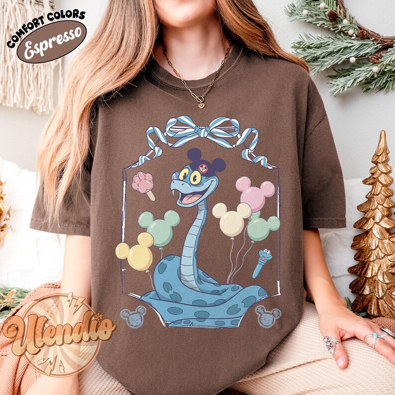 Puede incluir: Camiseta marr&oacute;n con una serpiente de dibujos animados con un sombrero de Mickey Mouse y globos. La serpiente es azul claro con manchas azul oscuro. La camiseta tiene las palabras "Comfort Colors" y "Espresso".