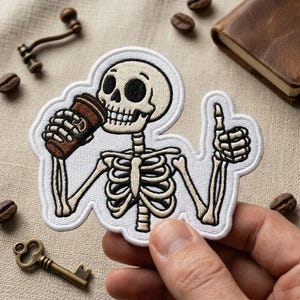 May include: An embroidered patch featuring a cream-colored skeleton with black outlines. The skeleton holds a brown coffee cup in one hand and gives a thumbs-up with the other. The design is outlined in white thread with a clean, finished border.