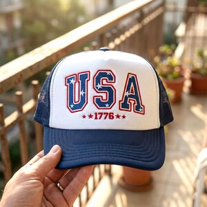 May include: A blue and white trucker hat with the word "USA" in red and blue lettering. The hat also features the year "1776" in red, with stars. The hat has a mesh back and a curved brim.