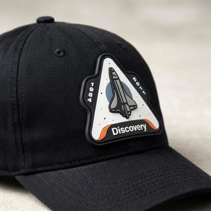 May include: A black baseball cap featuring a triangular patch on the front. The patch displays a space shuttle illustration, the text Discovery, and the years 1984 and 2011. The design includes a white background, black outlines, and orange accents.
