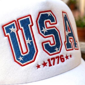 May include: White baseball cap with the word "USA" embroidered in blue with red trim and white stars. The year "1776" is embroidered in red below the letters. The cap is made of a soft, textured material.