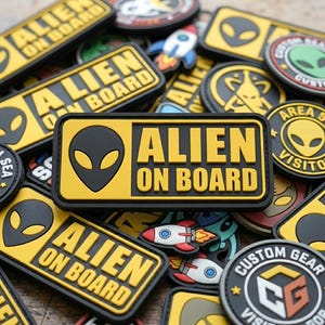 May include: A rectangular rubber patch featuring a yellow background with a black alien head icon and the text ALIEN ON BOARD in bold black letters. The patch is surrounded by various other alien-themed patches, including rocket ships and circular designs.