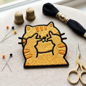 May include: An embroidered patch of a yellow tabby cat with dark stripes, shown making a hand gesture with both paws. The patch is surrounded by two brass thimbles, four sewing pins, a skein of black embroidery thread, and gold scissors.
