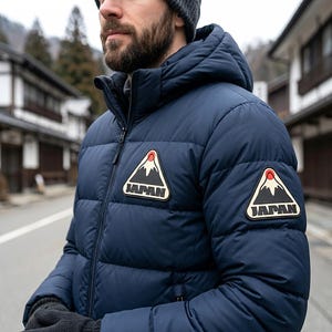 May include: A navy blue puffer jacket with a hood, featuring two triangular patches on the chest and sleeve. Each patch displays a black mountain graphic with a red sun above it and the word JAPAN printed in bold black capital letters below.