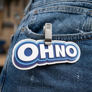 May include: A rectangular patch with rounded edges featuring the text OHNO in bold white letters. The text is layered over concentric blue and dark purple outlines. A silver metal clip is attached to the top center of the patch, securing it to blue denim fabric.