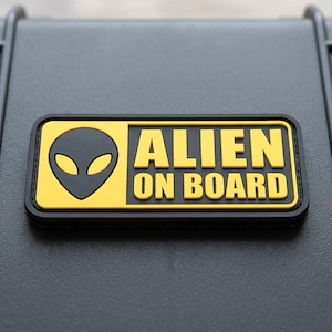 May include: A rectangular patch with a black border and a yellow background. On the left side is a black alien head icon. On the right side, the text ALIEN ON BOARD is printed in bold black capital letters. The patch has a raised, textured surface.