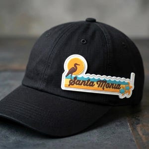 May include: A black baseball cap featuring a rectangular patch with a graphic of a bird, a yellow sun, blue waves, and the text Santa Monica in brown script. The patch includes a small blue turtle icon on the right side and a white border.