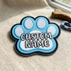 May include: A light blue embroidered paw print patch with the words "CUSTOM NAME" in white. The patch has a black border and is designed to be personalized. A dog bone and rope toy are in the background.