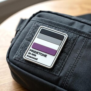 May include: A black tactical bag featuring a rectangular patch with a white border. The patch displays the asexual pride flag colors in horizontal stripes of black, gray, white, and purple, with the text PRIDETONE 28-1969 Asexuual printed below.