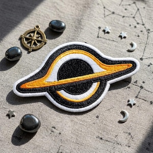 May include: Embroidered patch featuring a black hole design with white, yellow, and black stitching. The patch is surrounded by small silver stars, crescent moons, and polished black stones. A vintage brass compass is also present.