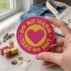 May include: A circular embroidered patch featuring a bright pink border and a yellow center with a pink heart. The text "DO NO HARM" is embroidered in yellow on the top half of the pink border, and "TAKE NO SHIT" is embroidered in yellow on the bottom.