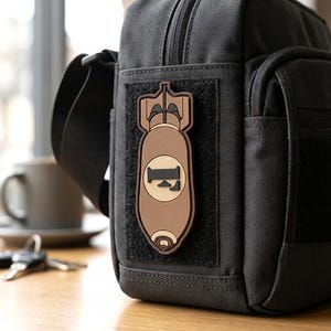 May include: A black tactical shoulder bag featuring a rectangular hook-and-loop panel on the front. Attached to the panel is a brown and tan rubberized patch shaped like an aerial bomb, displaying a stylized letter F in the center of the design.
