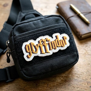 May include: A black tactical sling bag featuring a rectangular patch with the word "gryffindor" in a stylized yellow, red, and black font. The bag has a zippered main compartment, a front pocket with a hook-and-loop fastener, and an adjustable strap.