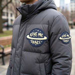 May include: A charcoal gray puffer jacket with a hood, featuring two circular navy blue and cream patches on the chest and sleeve. Each patch depicts a planet with rings and the text GIVE ME SPACE surrounded by small stars in a light yellow color.