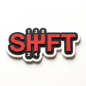May include: A decorative emblem featuring the word SHFT in bold red letters with a black outline and white border. The letter H is stylized as a manual transmission gear shift pattern with the numbers 1, 3, 5 on top and 2, 4 on the bottom.