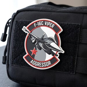 May include: A black tactical pouch featuring a circular PVC patch with a white, gray, and black F-16C Viper fighter jet graphic. The patch includes a red star, a red border, and the white text F-16C VIPER AGGRESSOR on a black background.