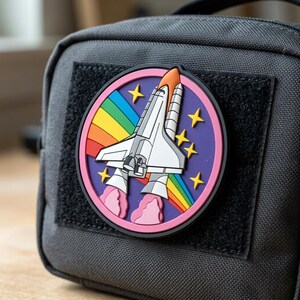May include: A dark gray fabric pouch featuring a circular rubber patch. The patch displays a white space shuttle launching against a purple background with a rainbow arc, yellow four-pointed stars, and pink exhaust clouds, all framed by a pink border.