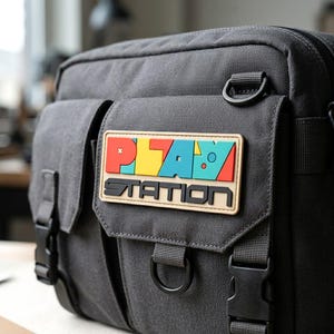 May include: A dark gray canvas messenger bag featuring a rectangular patch on the front flap. The patch displays the word PLAY in colorful block letters above the word STATION in black, bold, sans-serif text, with black plastic buckles.