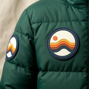 May include: A dark green quilted puffer jacket featuring two circular rubberized patches. Each patch displays a graphic of a stylized orange sun above layered wavy bands in shades of orange, brown, and dark blue against a cream background.