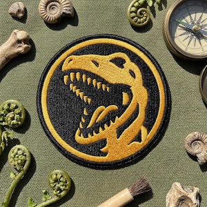 May include: Embroidered patch featuring a yellow Tyrannosaurus Rex head on a black circular background with a yellow border. The patch is surrounded by fossils, a compass, and a brush on a green fabric surface.