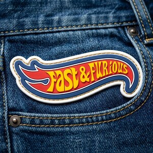 May include: A Fast & Furious logo patch featuring yellow lettering on a red background with a blue border and white outline, attached to blue denim fabric with visible gold stitching and a metal rivet near the pocket area.