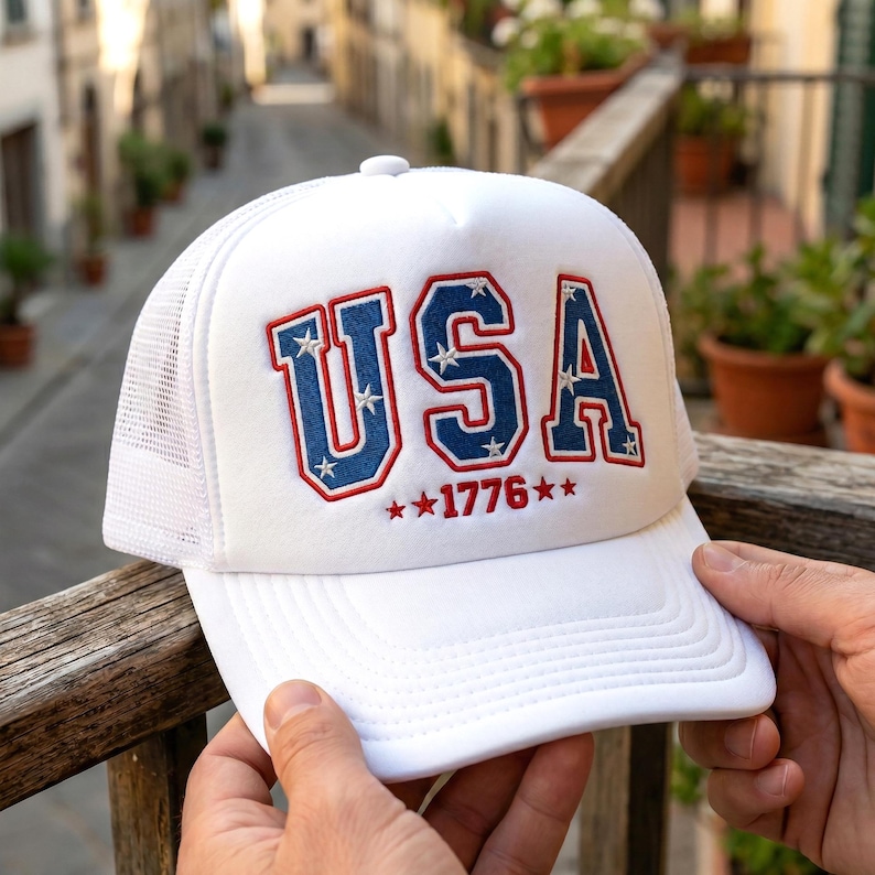 May include: White trucker hat with the word "USA" in red and blue lettering, with stars and the year "1776" embroidered on the front. The hat has a white mesh back and a curved brim.