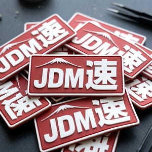 May include: A rectangular red patch with white raised lettering that reads JDM and the Japanese kanji character for speed. Above the text is a white graphic outline of Mount Fuji. The patch has a thick border and a matte finish.