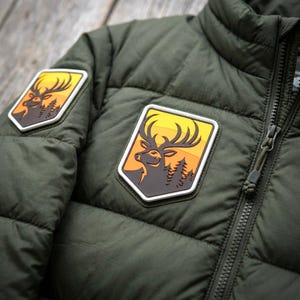 May include: An olive green quilted puffer jacket featuring two shield-shaped patches. Each patch displays a dark brown silhouette of a deer head with large antlers against a yellow and orange sunset background, accented by dark pine tree silhouettes.
